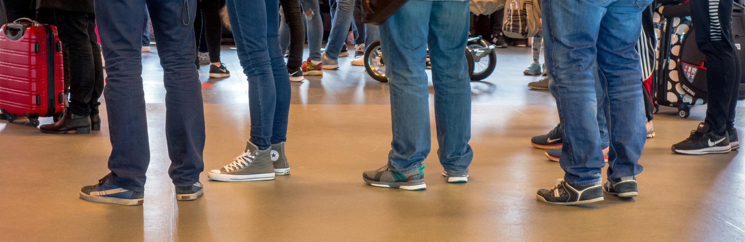 Une file d'attente vue au ras du sol : une rangée de jambes et de valises dans un hall.